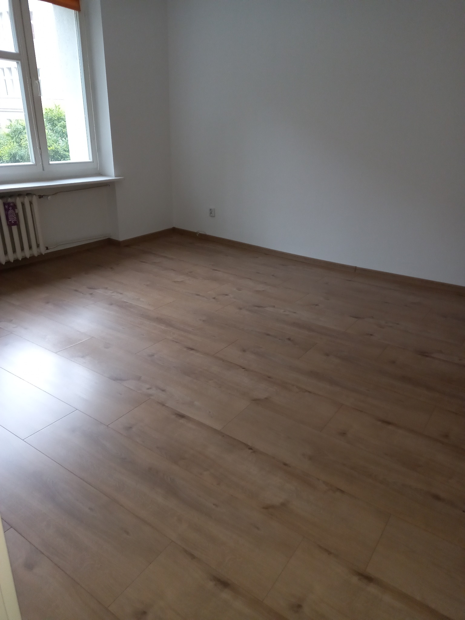 Pusty pokój z nowo ułożonymi panelami podłogowymi o naturalnym odcieniu drewna, białe ściany i okno z widokiem na zieleń za oknem, widoczny grzejnik pod oknem.