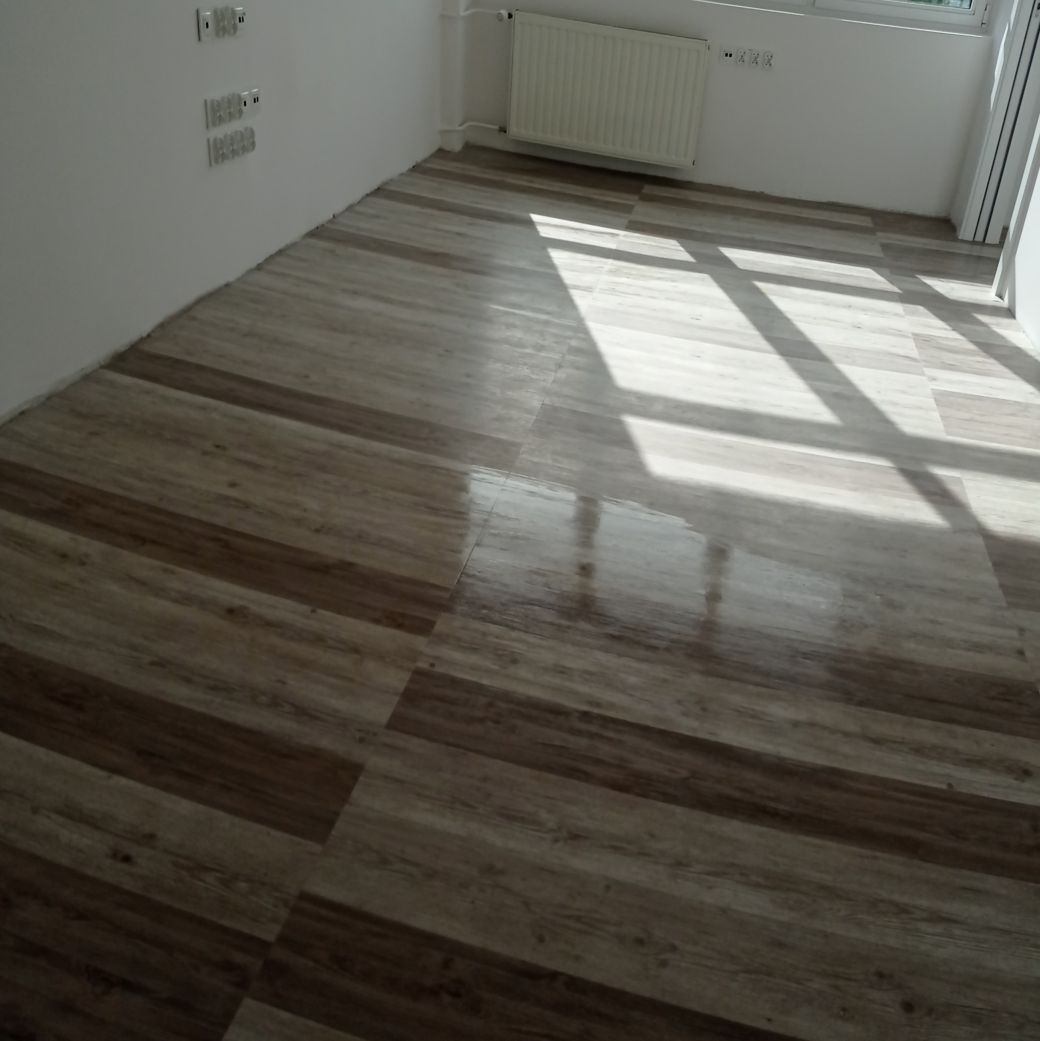 Ułożone panele podłogowe w nowym, jasnym pomieszczeniu z białymi ścianami, widoczne światło słoneczne padające przez okno.