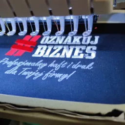 Proces tworzenia haftu z logo 'Oznakuj Biznes' na ciemnej tkaninie, widoczne igły maszyny hafciarskiej i kolorowe nici.
