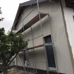 Elewacja domu w trakcie ocieplania styropianem, widoczne rusztowanie i zabezpieczone okno folią.