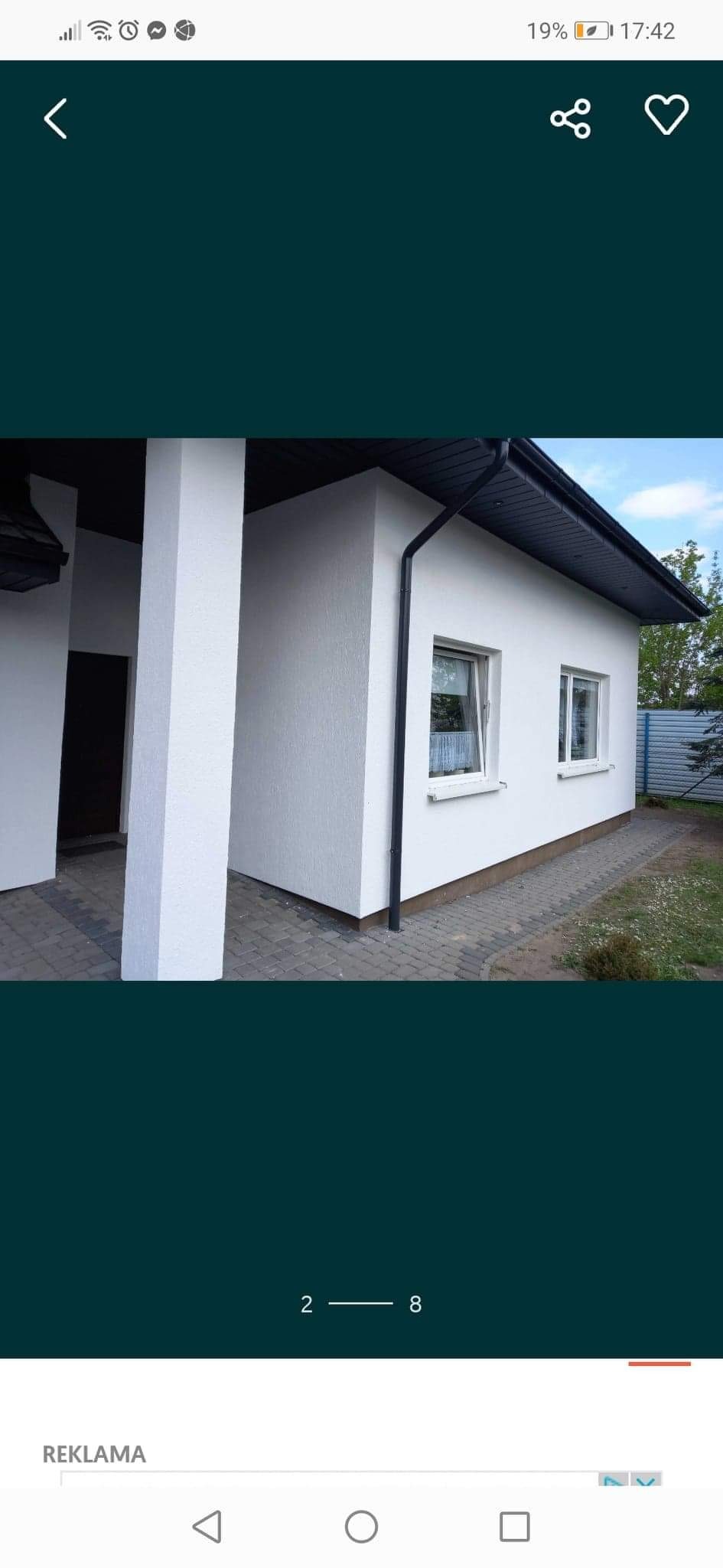 Biała elewacja budynku jednokondygnacyjnego z dwoma oknami, czarnym dachem i rynnami, widok z zewnątrz na kostkę brukową.