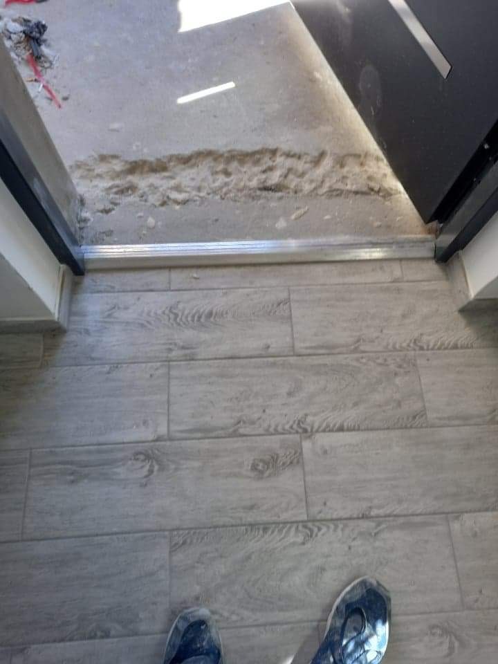 Fragment posadzki z szarych płytek imitujących drewno, widoczny próg drzwiowy z uszkodzonym podłożem i fragment butów osoby fotografującej.