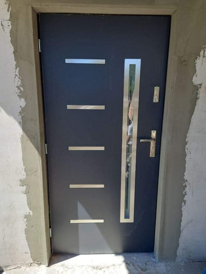 Nowoczesne, grafitowe drzwi wejściowe z czterema poziomymi, srebrnymi wstawkami i pionowym przeszkleniem w srebrnej ramie, osadzone w surowym, nieotynkowanym murze.