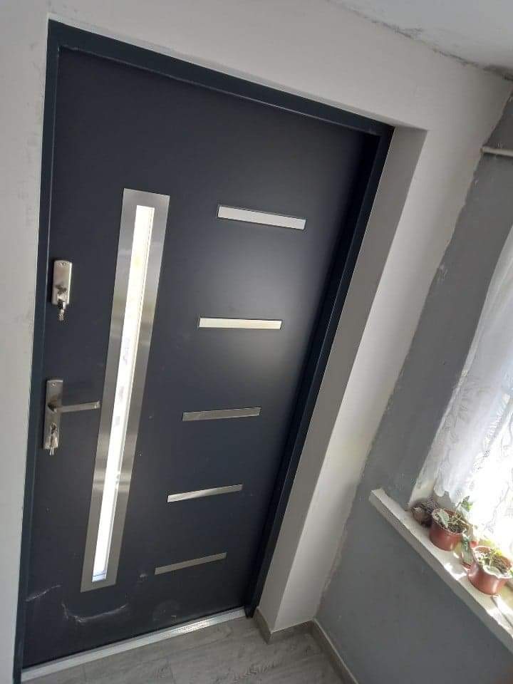 Ciemnoszare drzwi wejściowe z pionowym przeszkleniem i poziomymi, metalowymi akcentami, widoczne od strony wewnętrznej, z kluczem w zamku i doniczkami na parapecie w tle.
