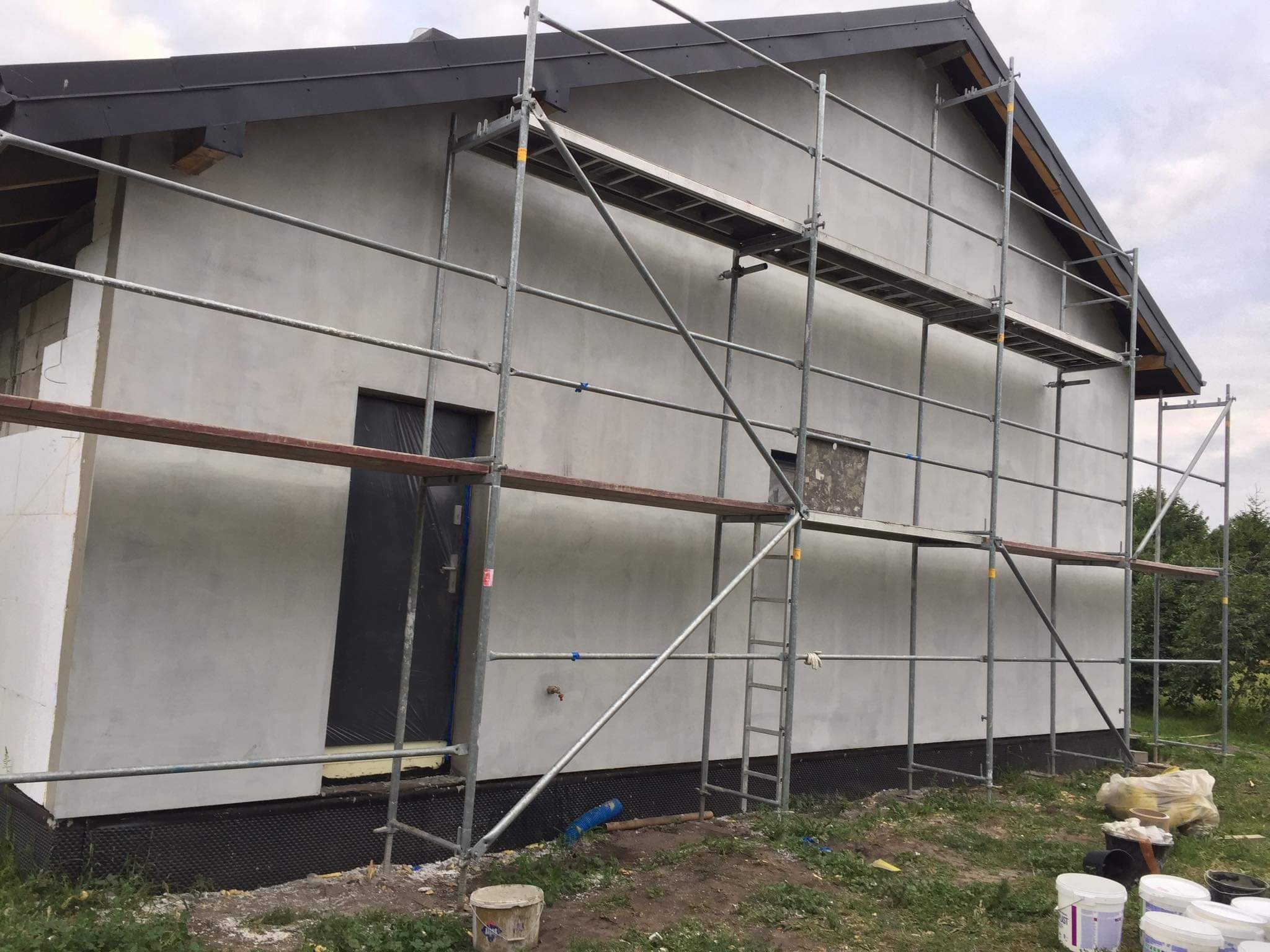 Elewacja budynku w trakcie prac wykończeniowych, widoczne rusztowanie, świeżo nałożona warstwa szarej gładzi, czarne wykończenie dachu, częściowo zakryte okno, oraz wiaderka z farbą i zaprawą u...