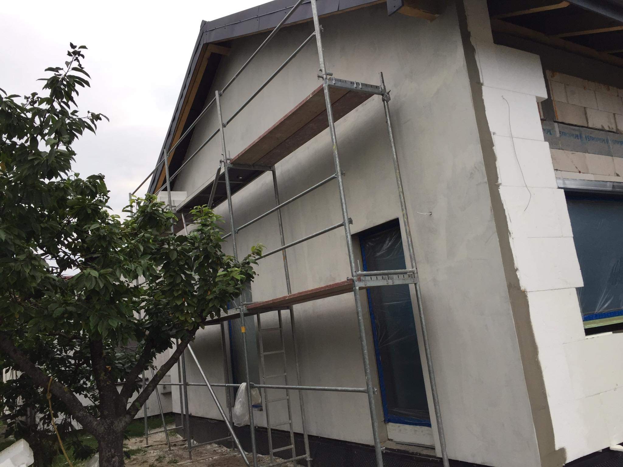 Elewacja domu w trakcie ocieplania styropianem, widoczne rusztowanie i zabezpieczone okno folią.