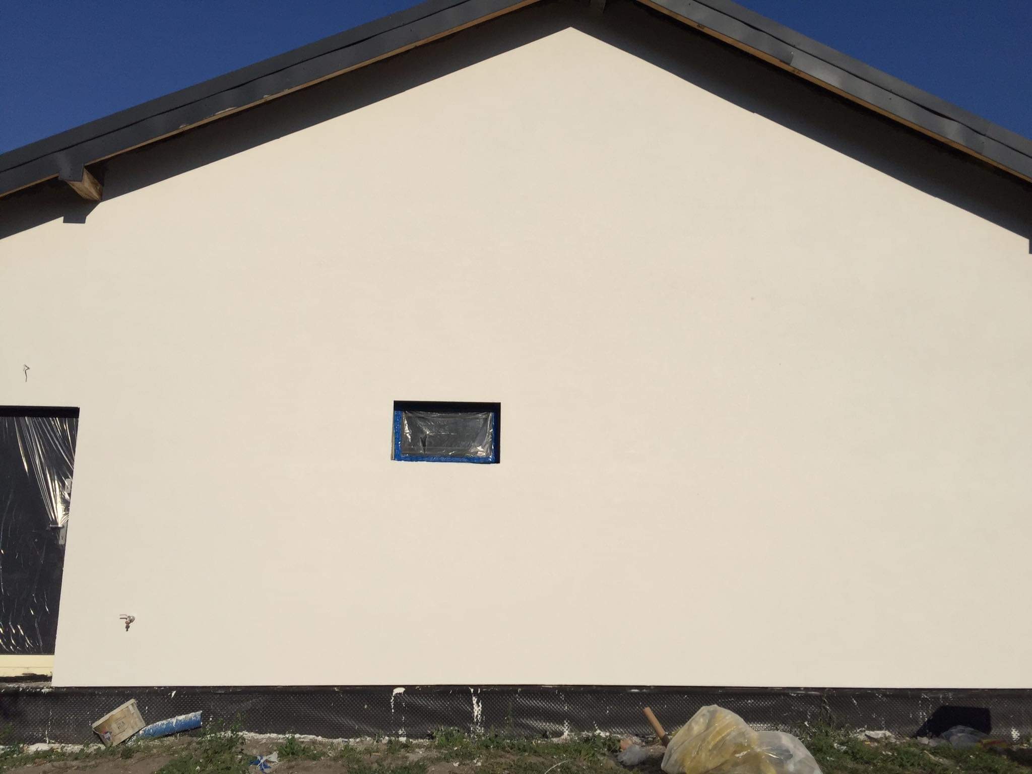Świeżo otynkowana ściana domu jednorodzinnego z zabezpieczonymi oknami folią malarską, widoczne ślady prac wykończeniowych w postaci resztek materiałów budowlanych u podstawy ściany.