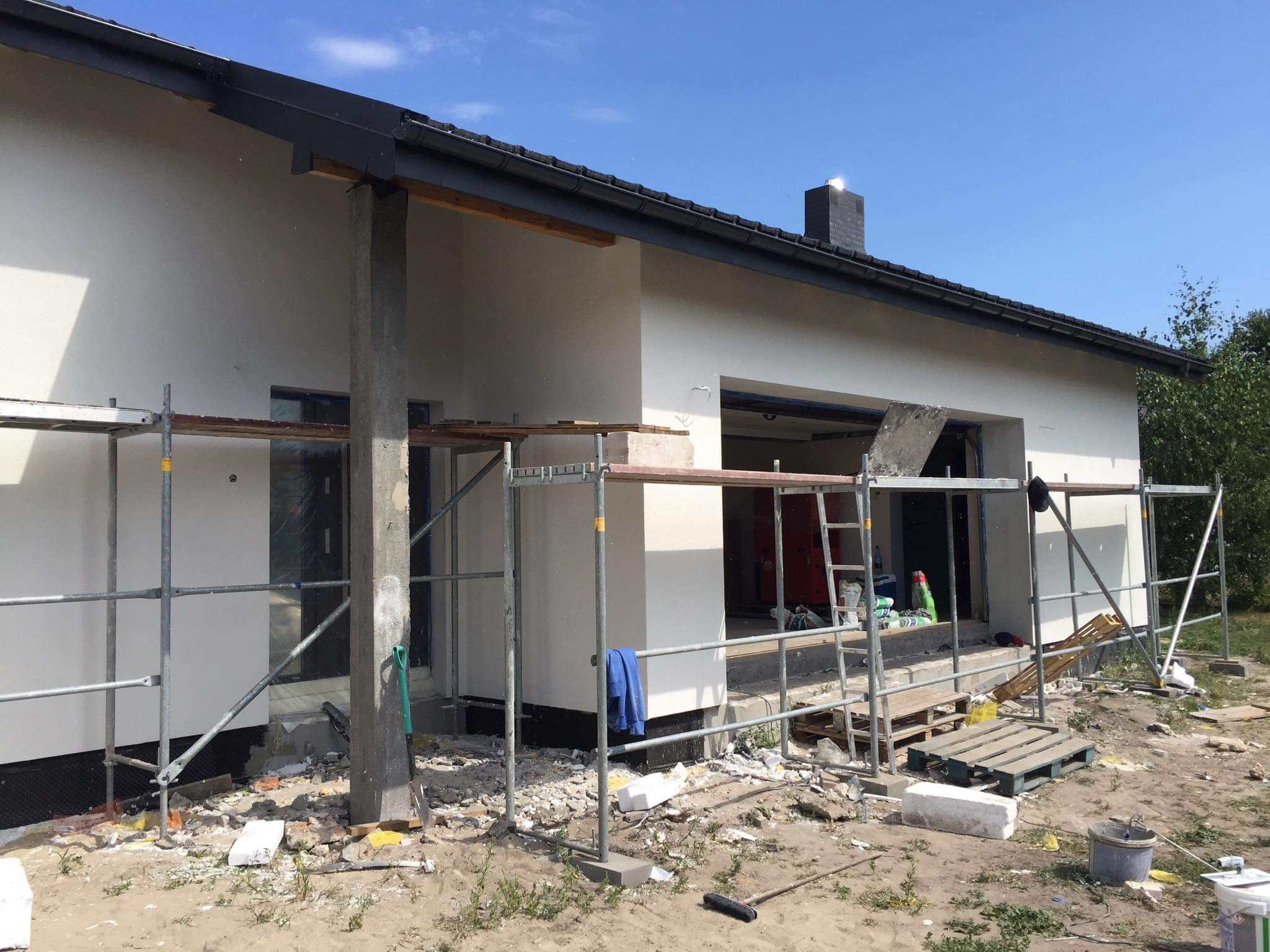 Nowo otynkowana elewacja domu jednorodzinnego w trakcie prac wykończeniowych, widoczne rusztowania, narzędzia i materiały budowlane, w tle błękitne niebo.