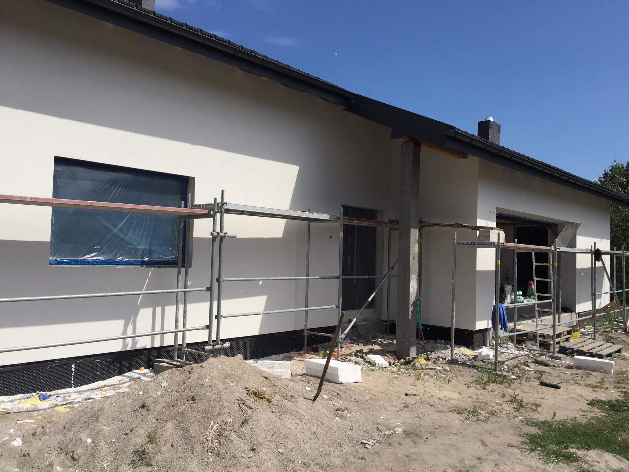 Elewacja domu jednorodzinnego w trakcie renowacji, widoczne rusztowania, zabezpieczone okno folią, świeżo nałożony tynk i przygotowany teren wokół budynku.