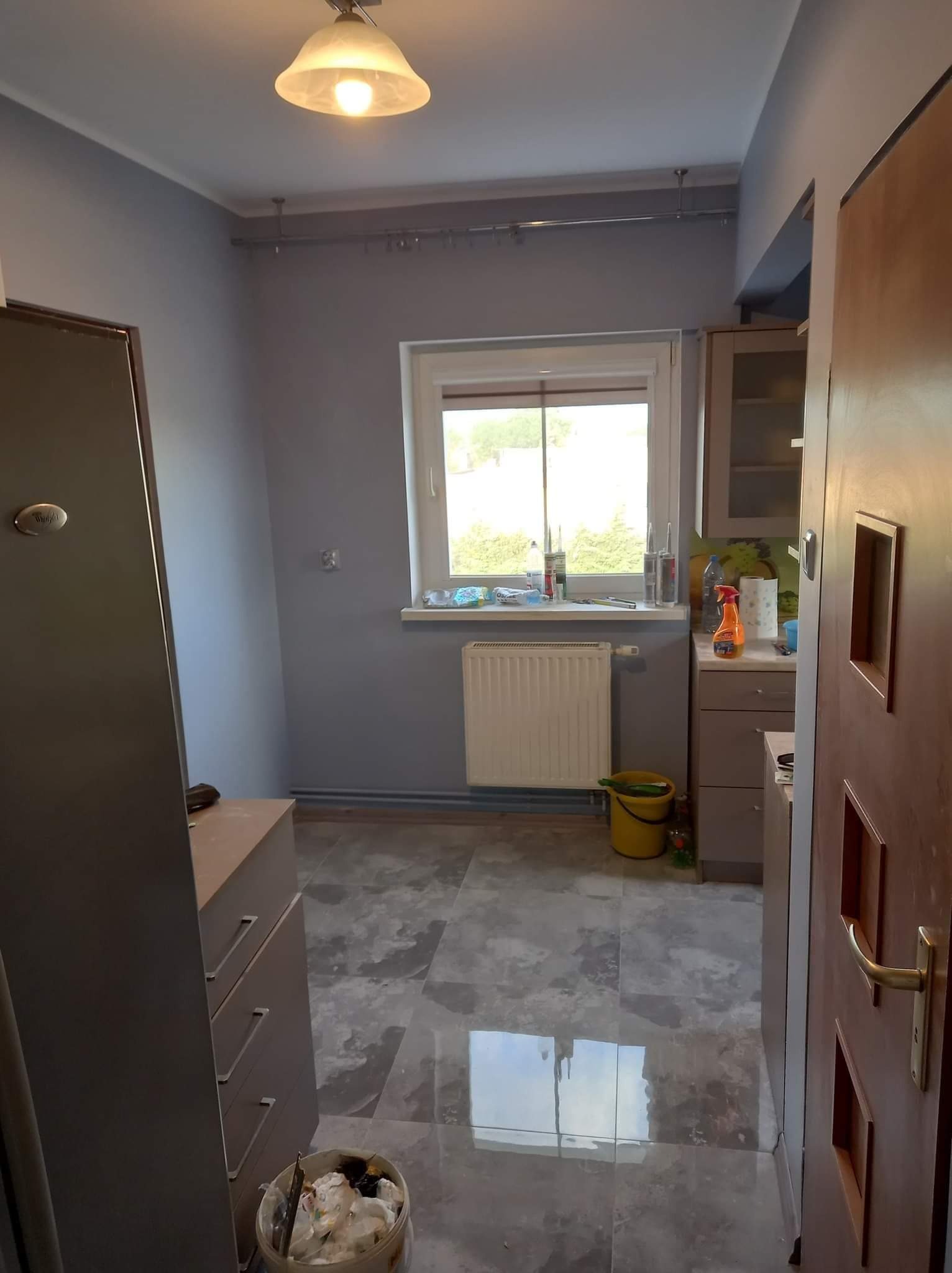Wnętrze kuchni w trakcie remontu, ściany pomalowane na szaro, widoczny grzejnik pod oknem, wiadro z farbą i narzędziami w rogu, marmurowe płytki na podłodze z odbiciem światła, częściowo widoczne...
