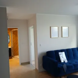 Salon po remoncie: widok na nową łazienkę z kabiną prysznicową, błyszczące płytki podłogowe, granatowa sofa z ulotkami i dwa puste obrazy na ścianie.