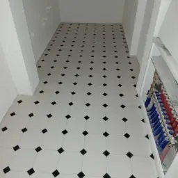 Biała podłoga wyłożona kwadratowymi płytkami z czarnymi rombami w wąskim korytarzu, widoczne rury instalacji grzewczej wzdłuż ściany po prawej stronie.