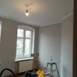 Pokój po malowaniu ścian na szaro, widoczne narzędzia malarskie, drabina z butelką wody, lampa budowlana i odkurzacz, surowe oświetlenie żarówki na suficie.