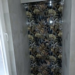 Remonty Volodymyr Muzyka - Wykończona kabina prysznicowa z baterią i deszczownicą, wyłożona płytkami z motywem roślinnym w odcieniach brązu, szarości i bieli, widok z boku na fragment ściany i okno.