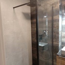 Remonty Volodymyr Muzyka - Nowoczesna kabina prysznicowa z czarną armaturą i szklaną ścianką w łazience wykończonej płytkami w dwóch odcieniach szarości i brązu. Widoczny fragment umywalki.