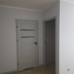 Remonty Volodymyr Muzyka - Szare drzwi wewnętrzne z przeszkleniami i ościeżnicą, widoczne na tle świeżo pomalowanej ściany i drewnopodobnej podłogi. Widoczny fragment drugiego pomieszczenia.