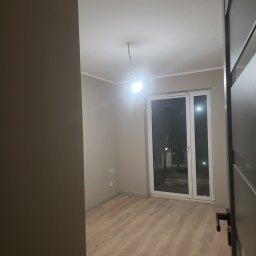 Remonty Volodymyr Muzyka - Jasne panele podłogowe w nowym pokoju z szarymi ścianami i białymi oknami balkonowymi. Widok zza uchylonych, ciemnych drzwi.