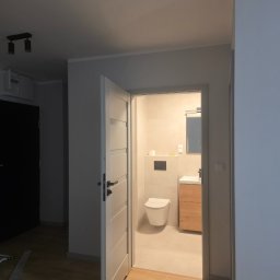 Remonty Volodymyr Muzyka - Otwarta biała stolarka drzwiowa ukazuje wykończoną, jasną łazienkę z podwieszaną toaletą, drewnianą szafką pod umywalką i lustrem. Widok z korytarza z drewnianą podłogą.