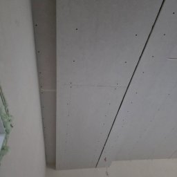 Remonty Volodymyr Muzyka - Montaż płyt gipsowo-kartonowych na suficie, widoczne łączenia i śruby mocujące. W tle fragment ściany i wisząca żarówka.
