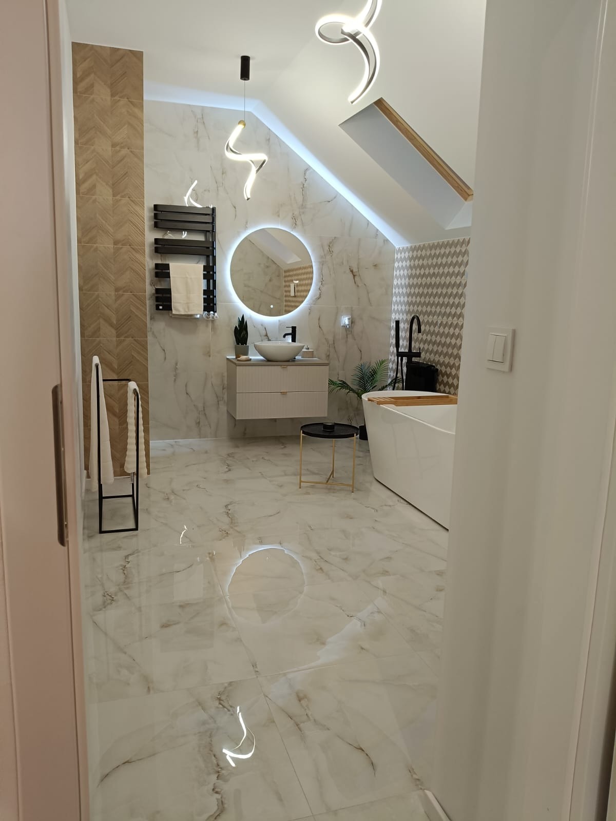 Nowoczesna łazienka na poddaszu z marmurową podłogą i ścianami, okrągłym lustrem z podświetleniem LED, wolnostojącą wanną z czarną armaturą i designerskim oświetleniem.