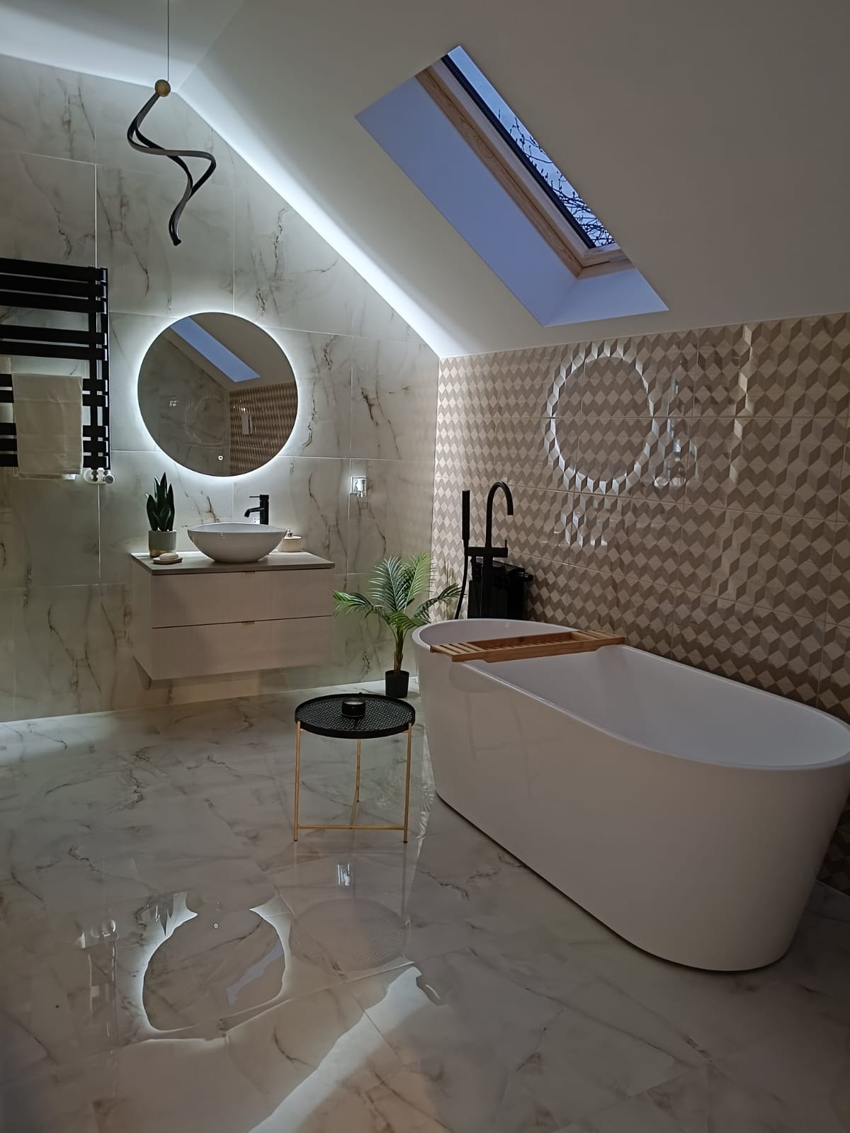 Nowoczesna łazienka na poddaszu z białą wanną wolnostojącą, umywalką na szafce z okrągłym lustrem LED, marmurową okładziną ścian i podłogi oraz oknem dachowym.