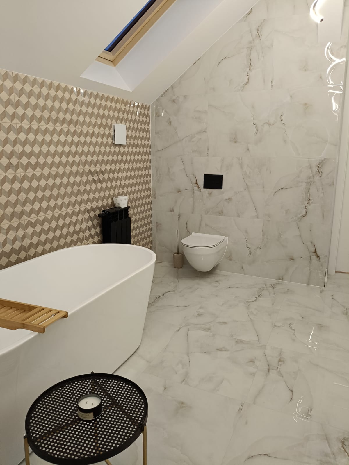 Nowoczesna łazienka na poddaszu z wolnostojącą białą wanną, geometryczną mozaiką na ścianie i marmurowymi płytkami, oświetlona przez okno dachowe.