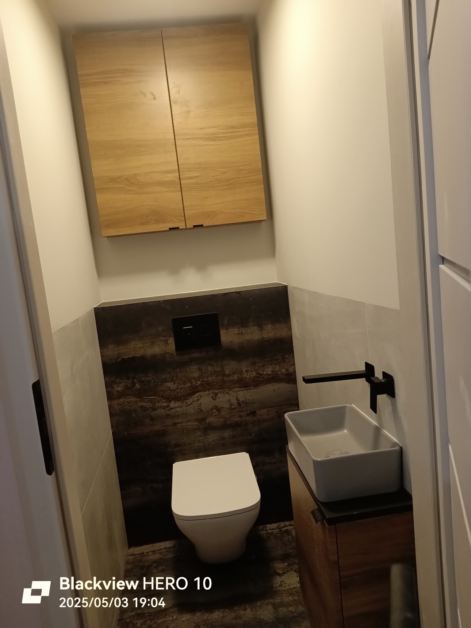 Nowoczesna toaleta z podwieszaną miską WC, umywalką nablatową na drewnianej szafce i szafką wiszącą. Ciemne płytki imitujące kamień kontrastują z bielą ścian.