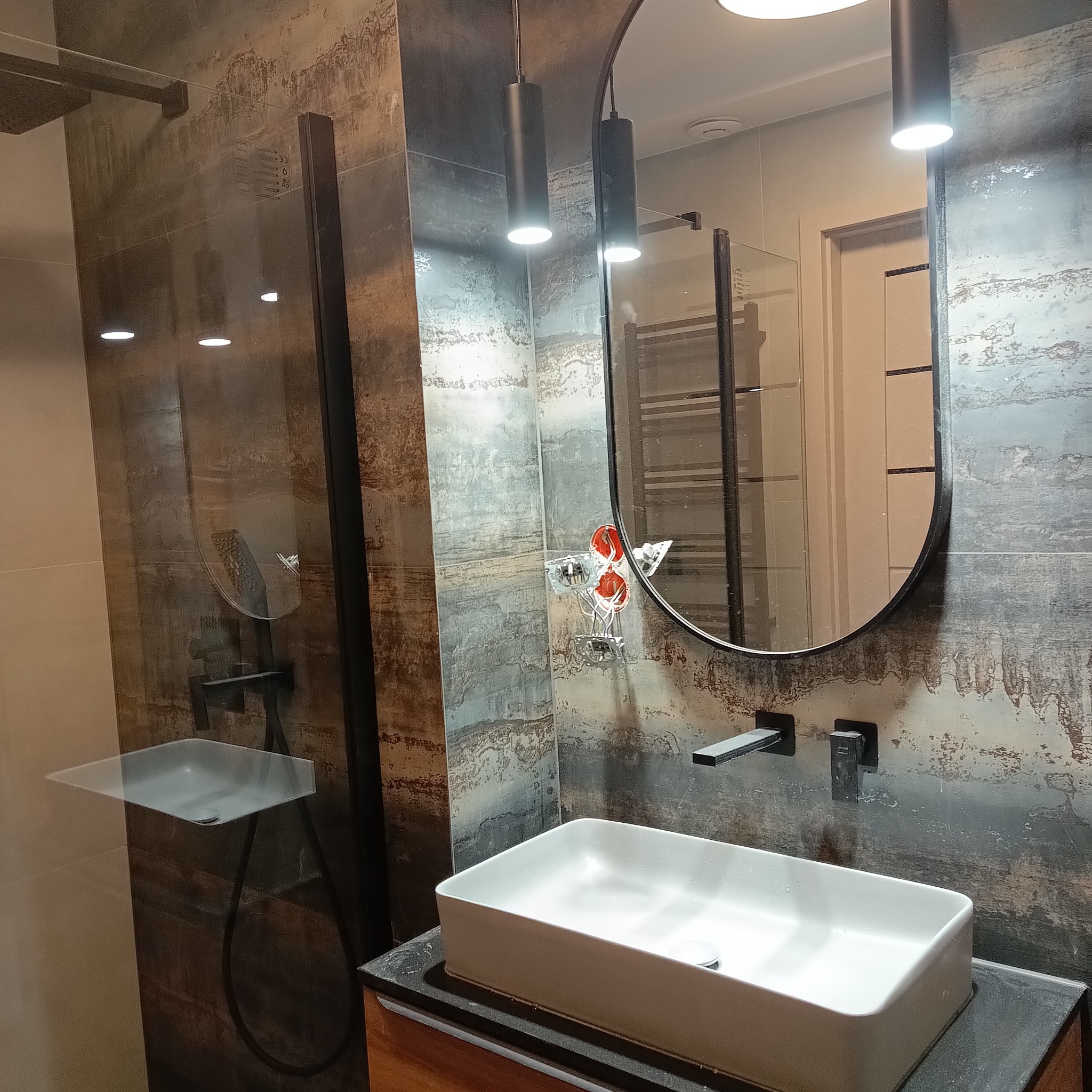 Nowoczesna łazienka z umywalką nablatową, armaturą w kolorze czarnym i lustrem owalnym. Na ścianach płytki imitujące rdzę. Widoczne niedokończone gniazdka elektryczne.