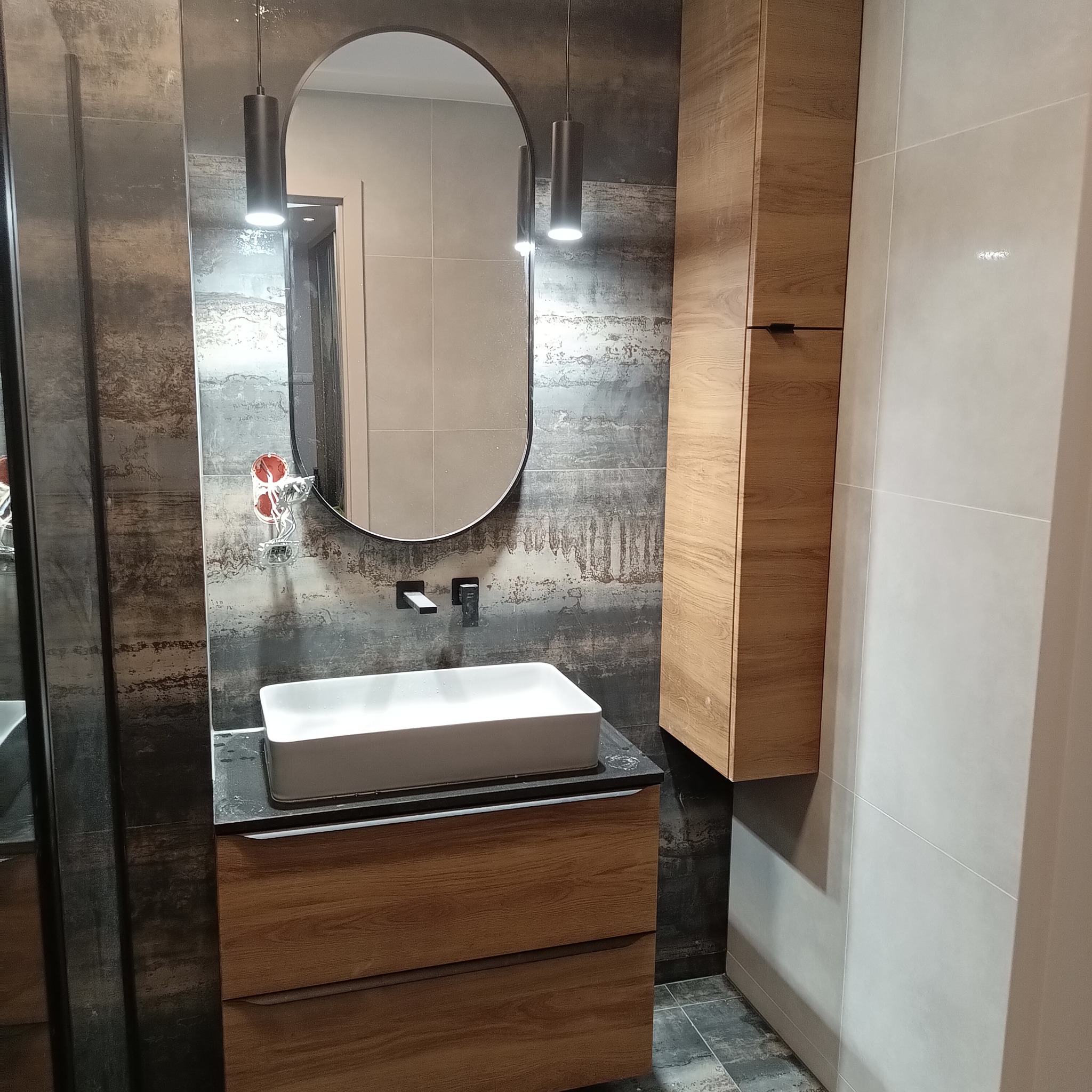 Nowoczesna łazienka z owalnym lustrem, białą umywalką nablatową i drewnianą szafką. Ściany wyłożone płytkami imitującymi beton i jasnymi płytkami. Oświetlenie punktowe.