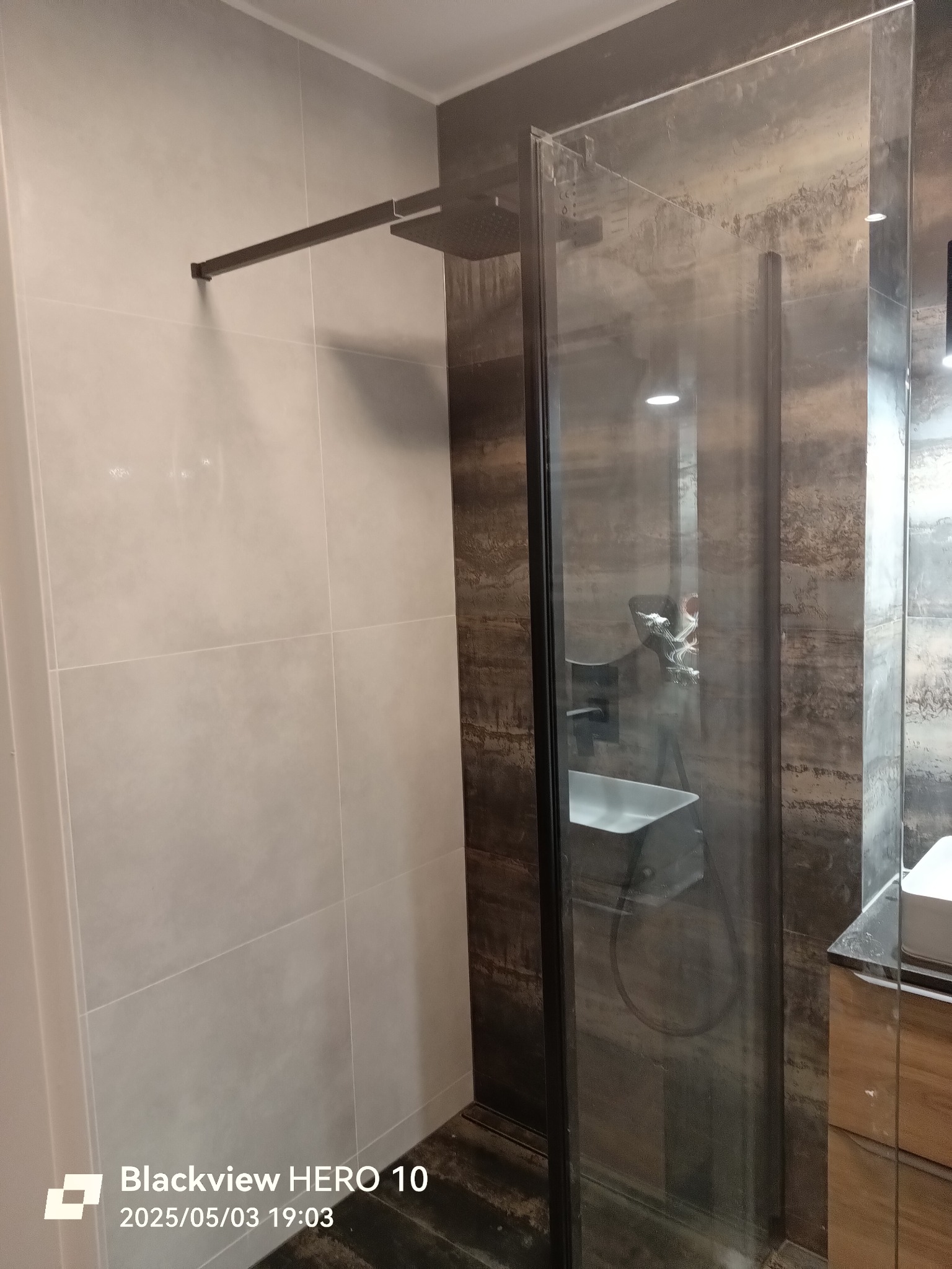 Nowoczesna kabina prysznicowa z czarną armaturą i szklaną ścianką w łazience wykończonej płytkami w dwóch odcieniach szarości i brązu. Widoczny fragment umywalki.