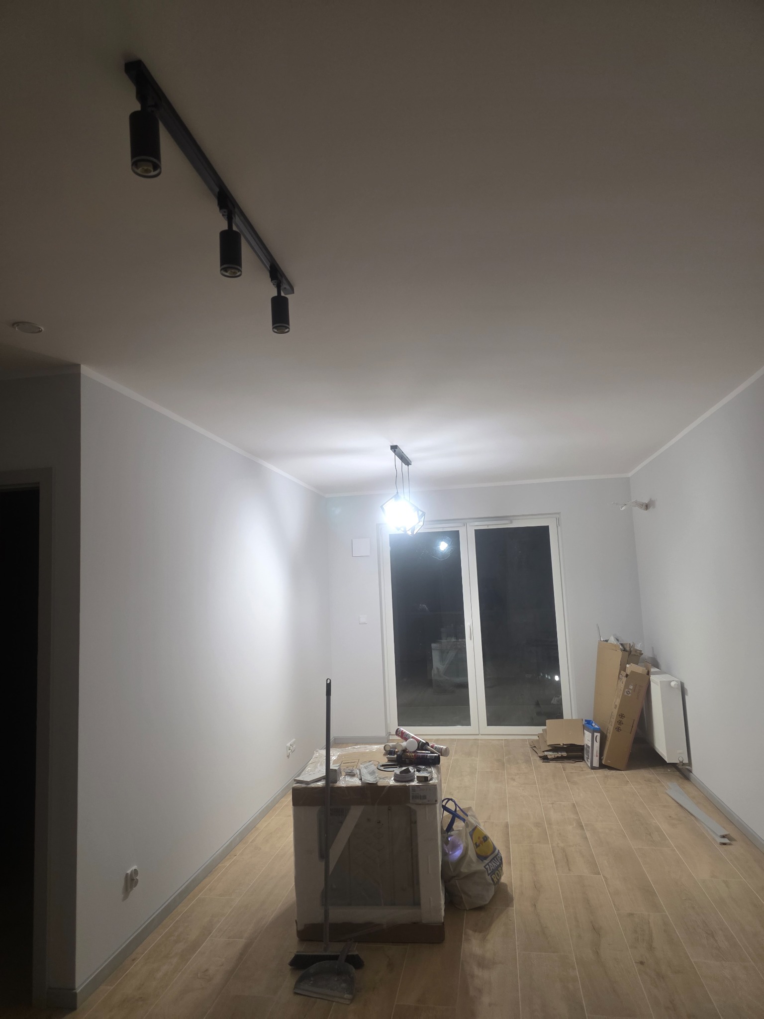 Jasne wnętrze po remoncie z drewnianą podłogą, białymi ścianami i nowoczesnym oświetleniem szynowym. Widoczne narzędzia i materiały budowlane sugerują etap wykończeniowy.