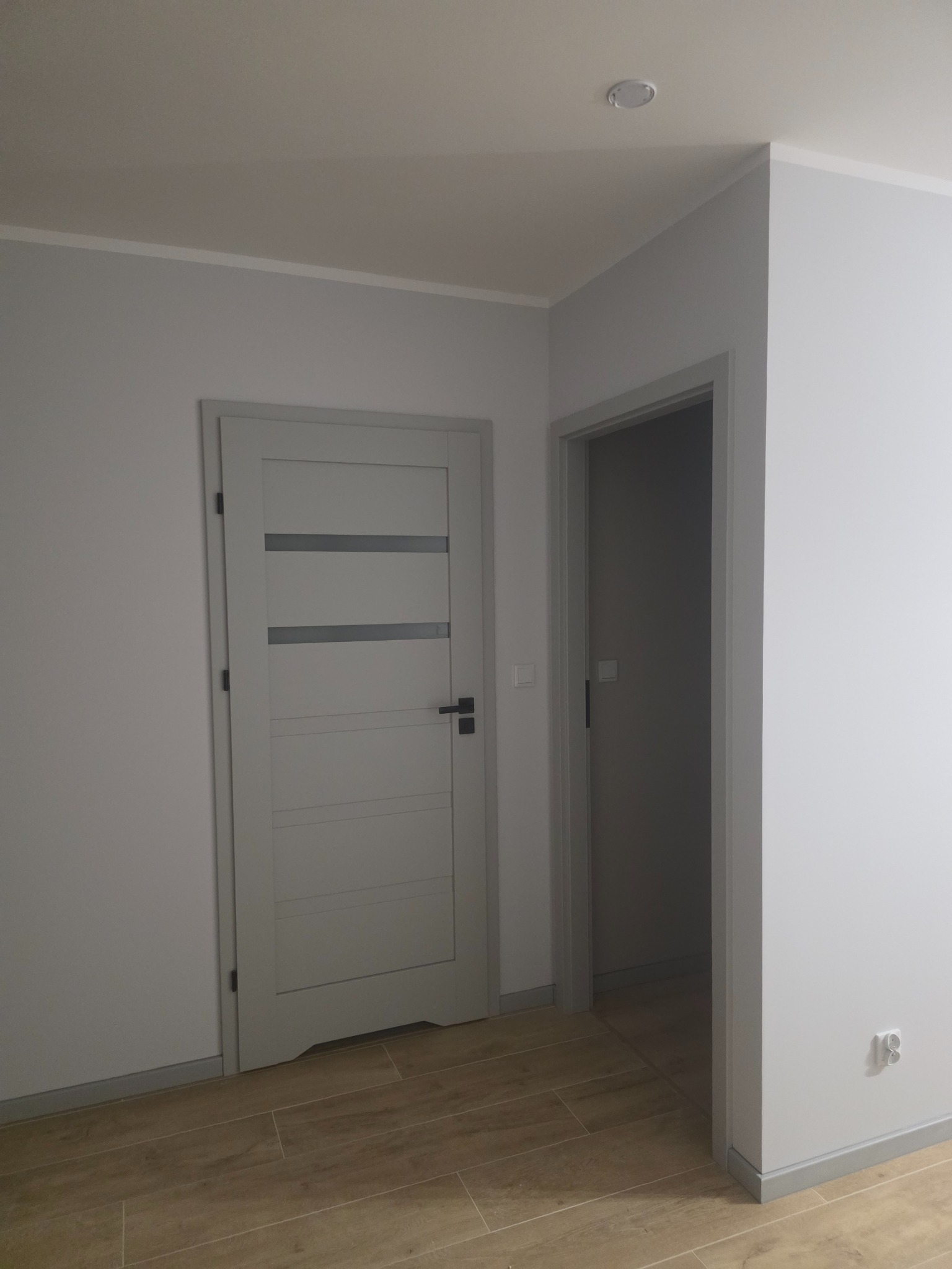 Szare drzwi wewnętrzne z przeszkleniami i ościeżnicą, widoczne na tle świeżo pomalowanej ściany i drewnopodobnej podłogi. Widoczny fragment drugiego pomieszczenia.