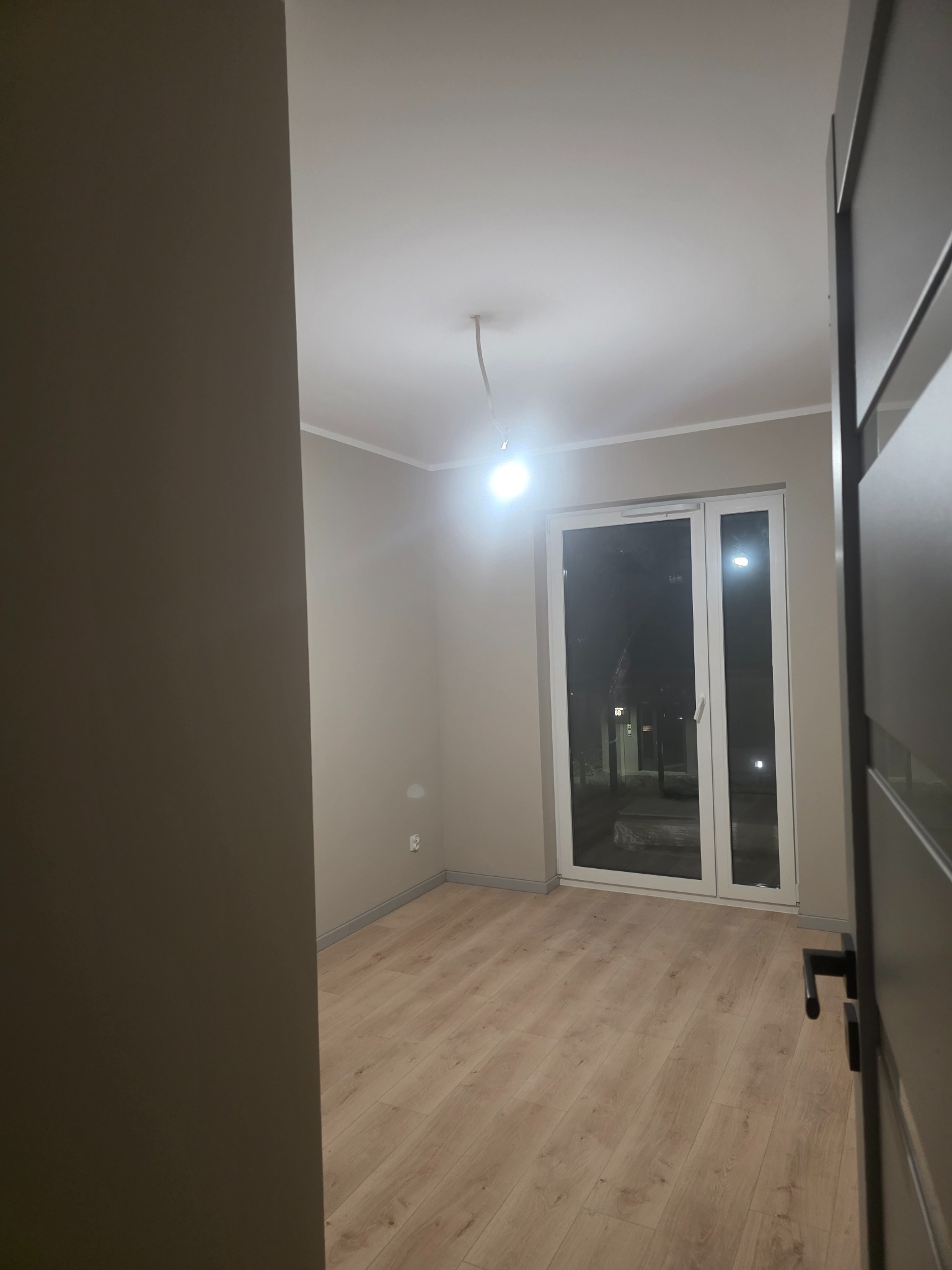 Jasne panele podłogowe w nowym pokoju z szarymi ścianami i białymi oknami balkonowymi. Widok zza uchylonych, ciemnych drzwi.