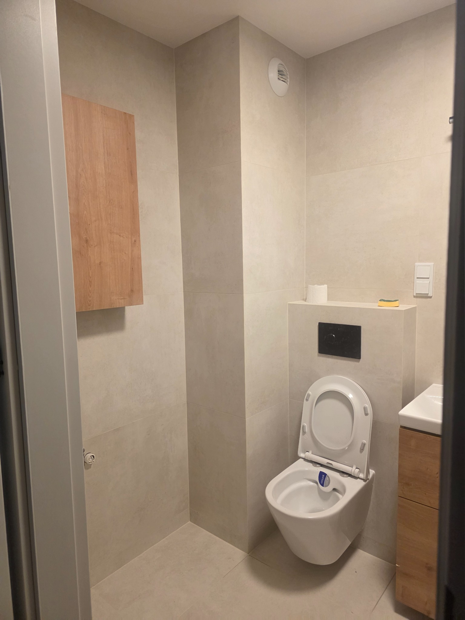 Jasna łazienka po remoncie z podwieszaną toaletą, szafką nad nią i umywalką z szafką. Minimalistyczny design, beżowe płytki, widoczny fragment drzwi.