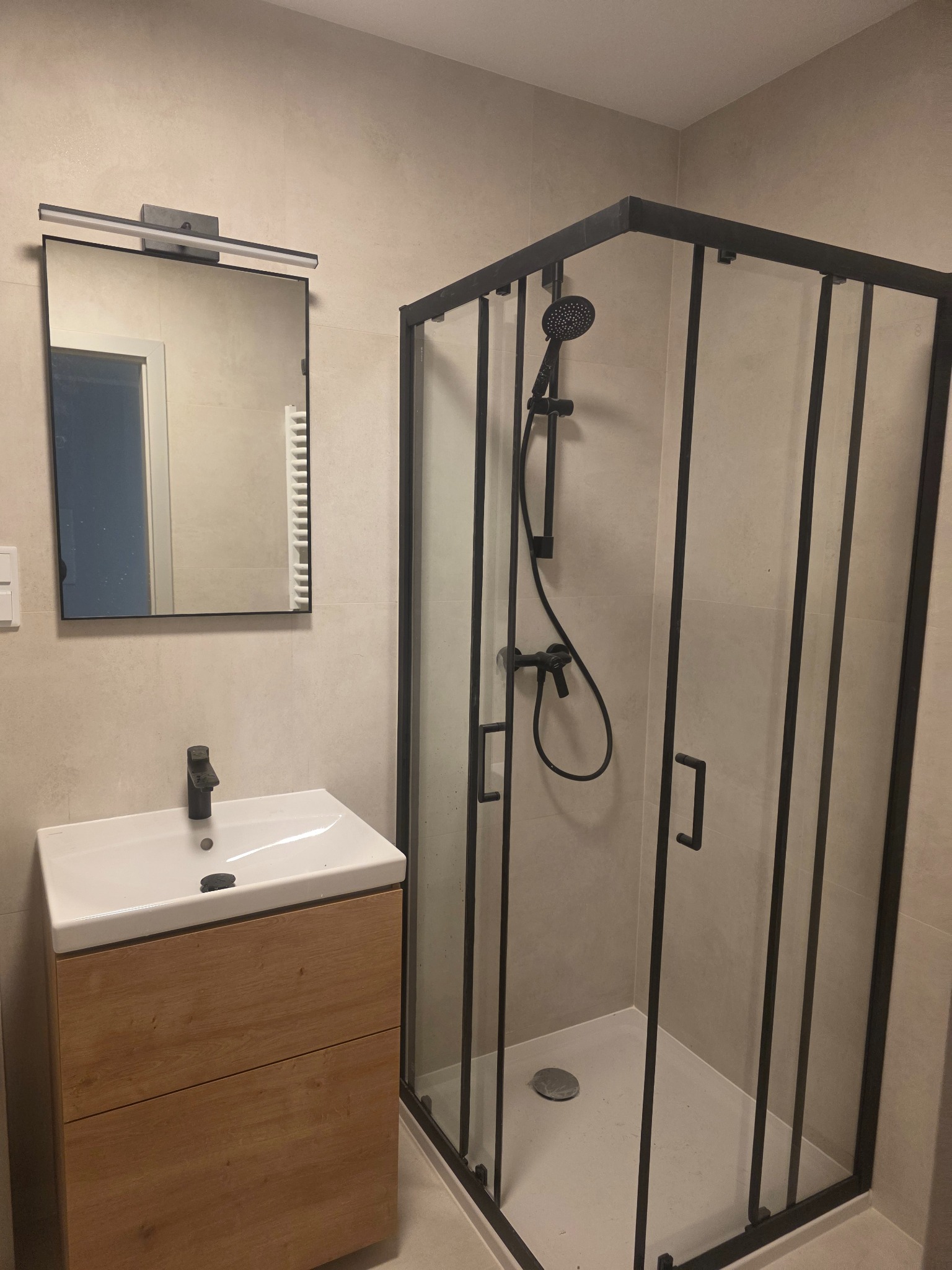 Nowoczesna łazienka z kabiną prysznicową w czarnej ramie, umywalką z drewnianą szafką i lustrem z oświetleniem. Beżowe płytki na ścianach tworzą spójną całość.