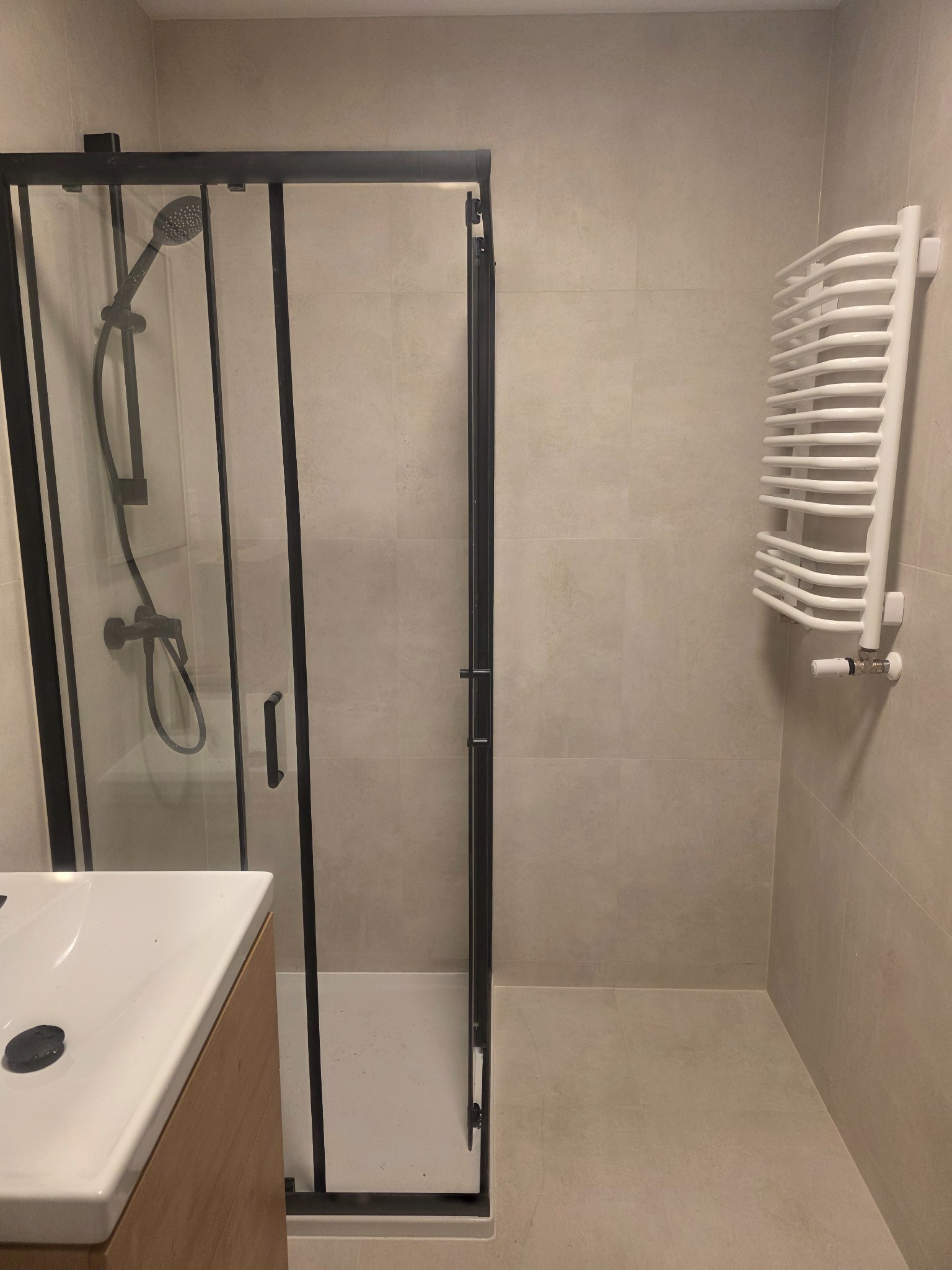 Nowoczesna łazienka z kabiną prysznicową typu walk-in z czarnymi profilami, grzejnikiem drabinkowym i umywalką nablatową na drewnianej szafce. Minimalistyczny design.