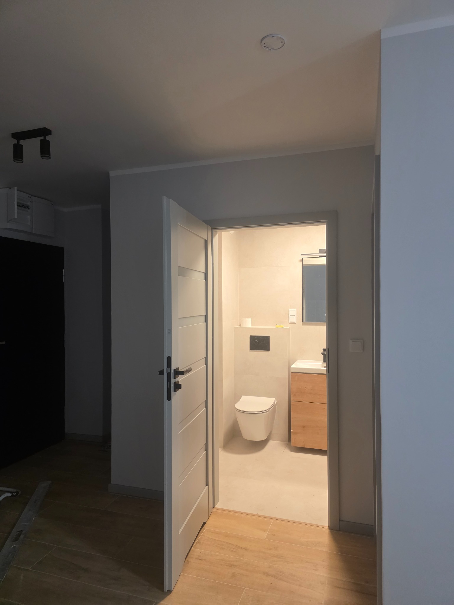 Otwarta biała stolarka drzwiowa ukazuje wykończoną, jasną łazienkę z podwieszaną toaletą, drewnianą szafką pod umywalką i lustrem. Widok z korytarza z drewnianą podłogą.