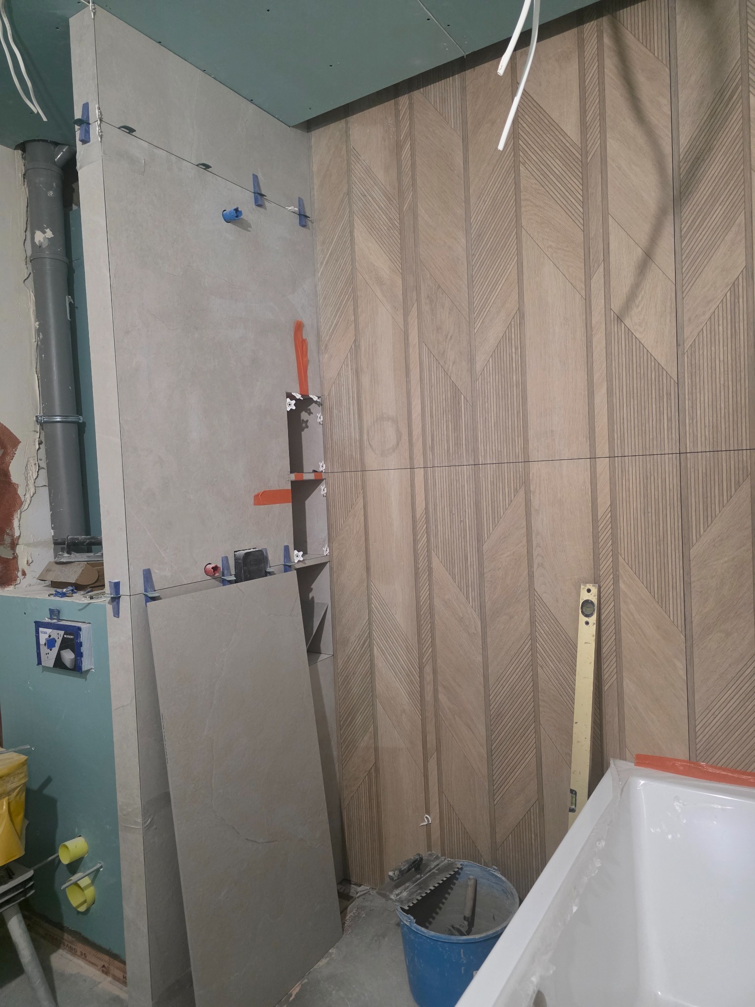 Układanie płytek z wzorem jodełki na ścianie łazienki, widoczne narzędzia glazurnicze i poziomica. Prace wykończeniowe w toku, z użyciem systemu poziomowania płytek.