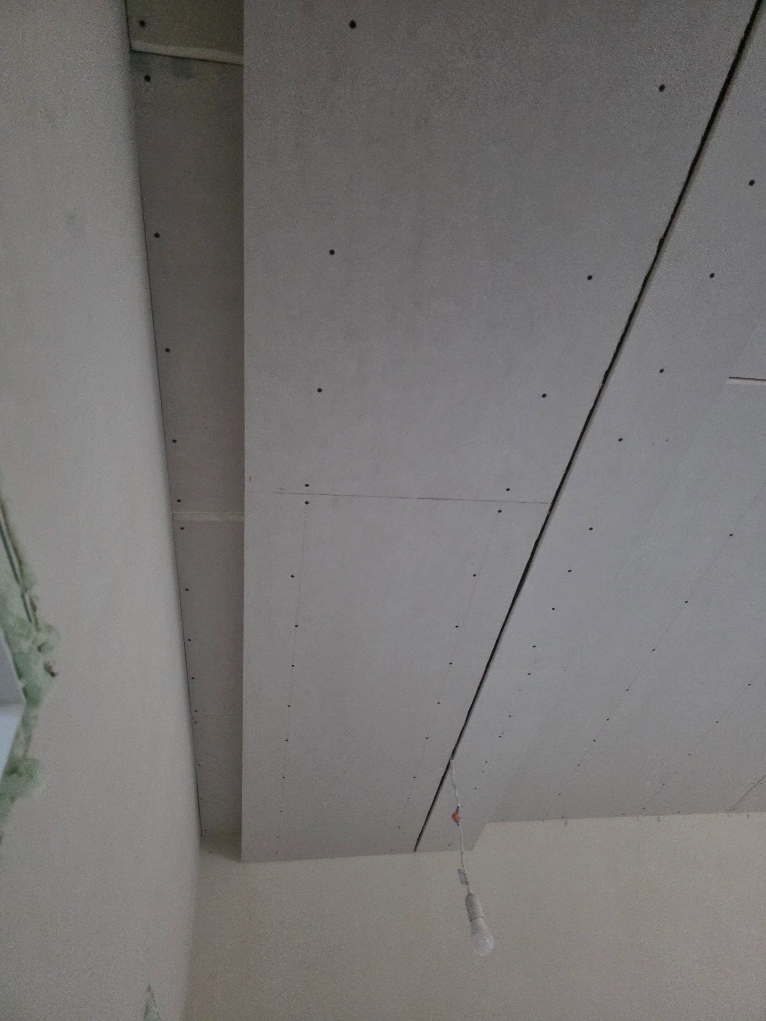 Montaż płyt gipsowo-kartonowych na suficie, widoczne łączenia i śruby mocujące. W tle fragment ściany i wisząca żarówka.