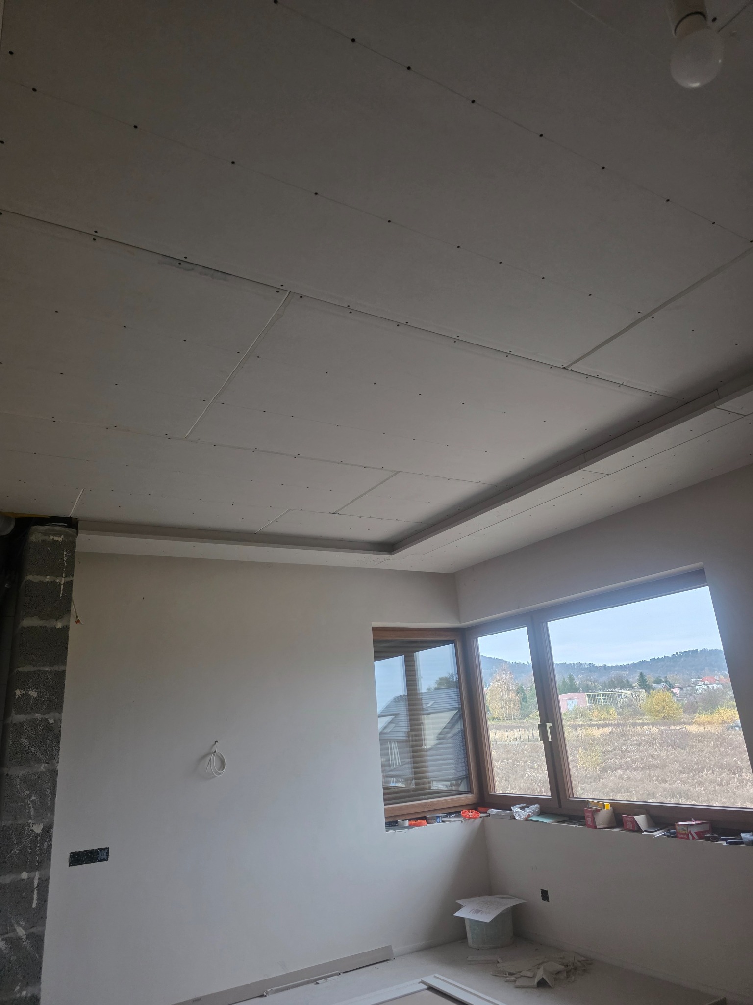 Sufit z płyt kartonowo-gipsowych w trakcie montażu, widoczne łączenia i śruby, surowy stan wykończenia wnętrza z oknami i widokiem na zewnątrz.