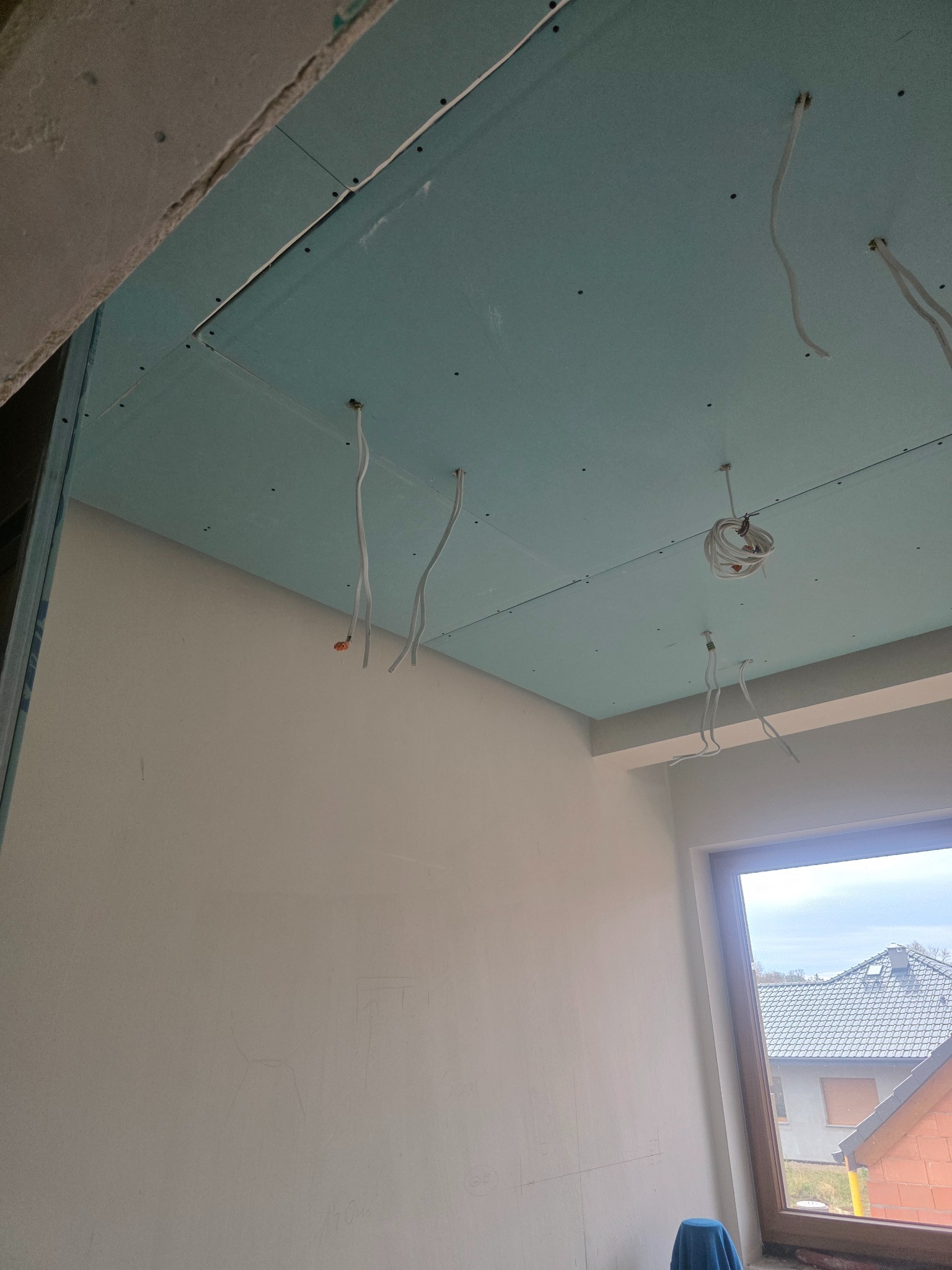 Montaż sufitu podwieszanego z płyt gipsowo-kartonowych, widoczne kable elektryczne. Ściana przygotowana do dalszych prac wykończeniowych, okno w tle.