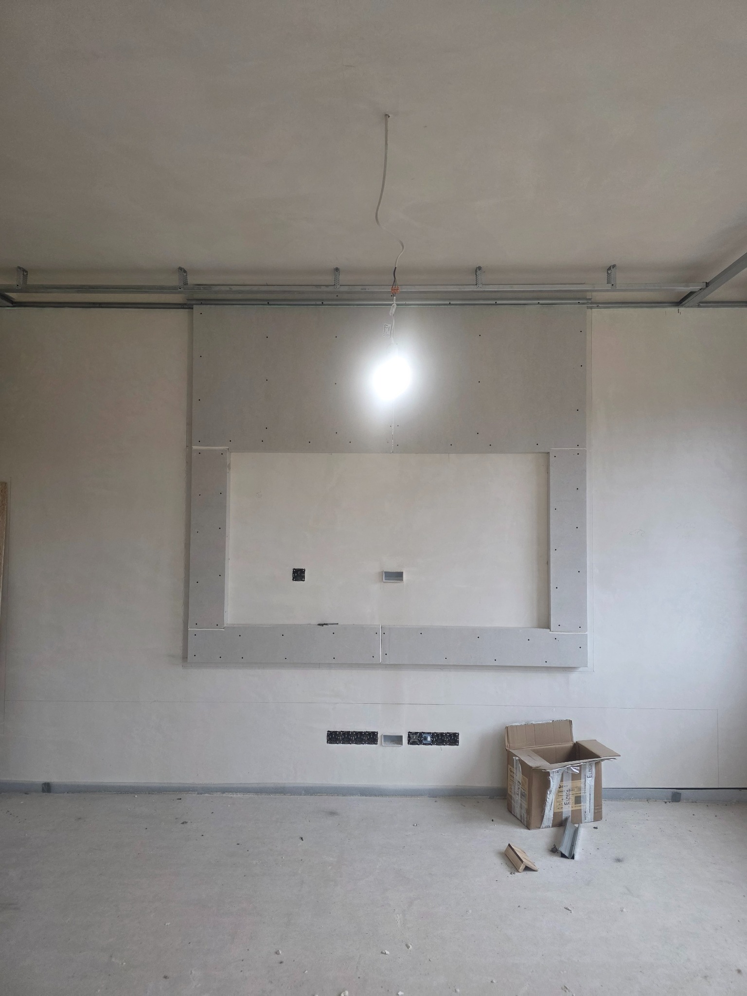 Ściana z płyt kartonowo-gipsowych w trakcie montażu, widoczne profile i otwory na instalacje. Surowy stan wykończenia, karton i narzędzia na podłodze.