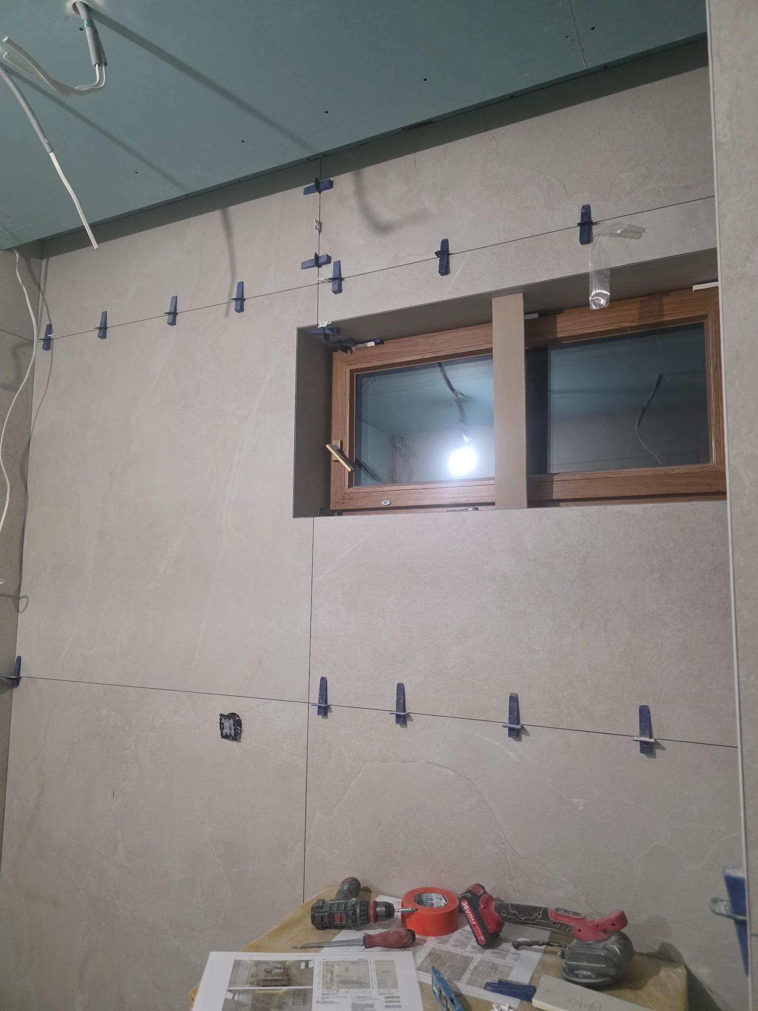 Ściana z beżowymi płytkami, okno z drewnianą ramą, niebieskie klipsy poziomujące, narzędzia na stole. Etap prac wykończeniowych w łazience.