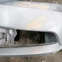 Szary zdemontowany zderzak samochodowy z widocznymi miejscami wypełnionymi szpachlą przed lakierowaniem, ukazujący proces naprawy karoserii.