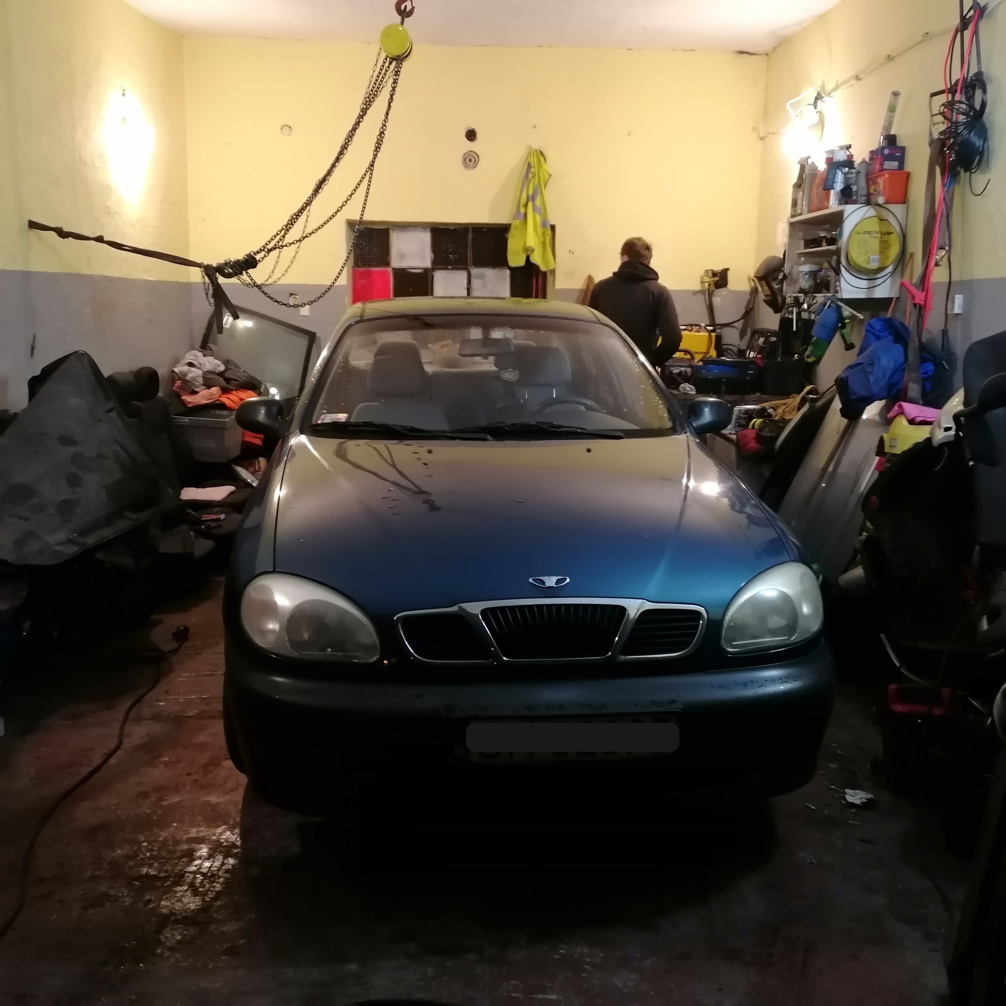 Niebieski samochód osobowy w warsztacie, podwieszony łańcuch z hakiem suwnicy nad maską, w tle widoczne regały z narzędziami i pracownik.