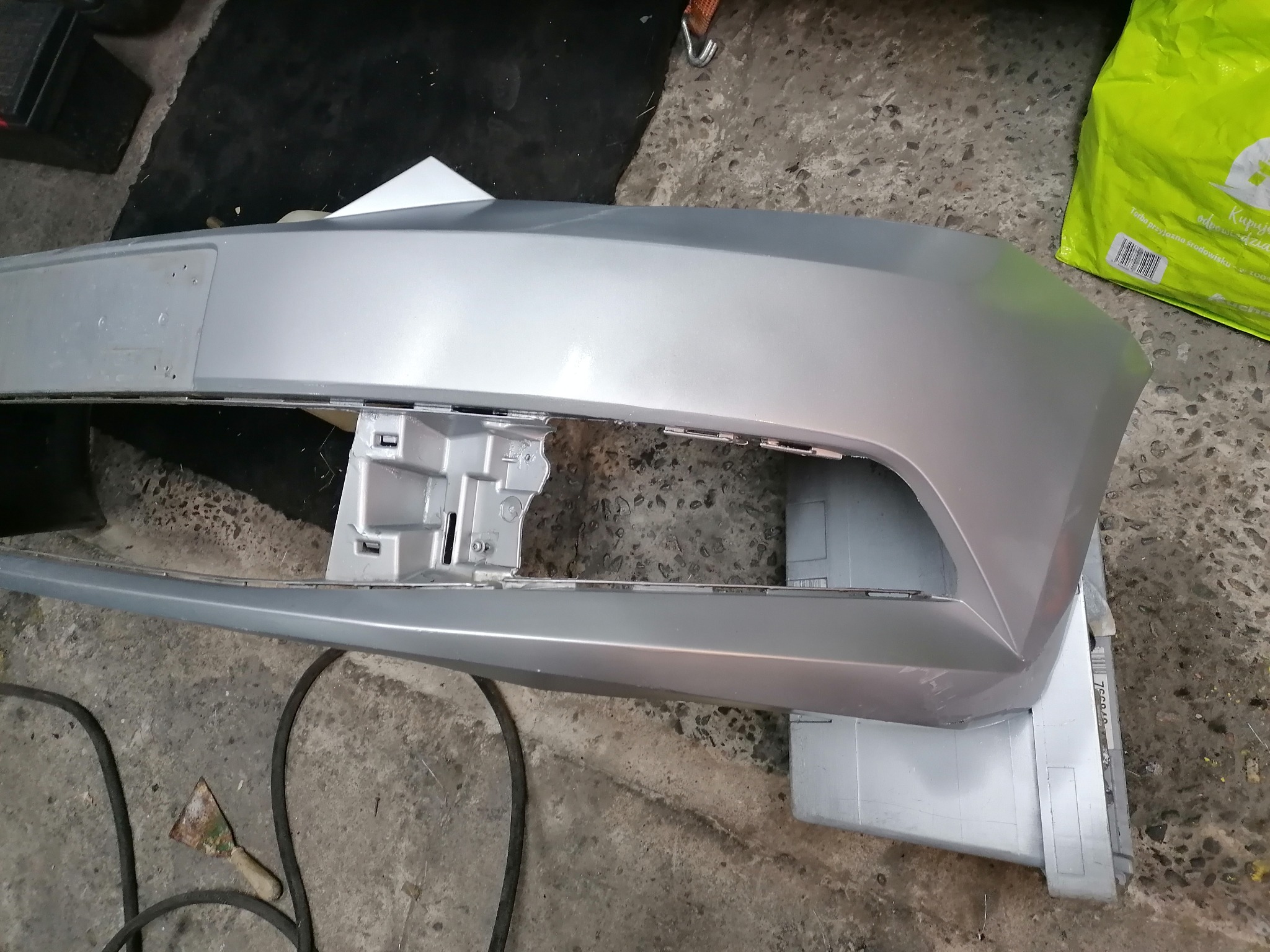 Srebrny, surowy zderzak samochodowy, przygotowany do lakierowania, leży na szarym podłożu warsztatowym, widoczne ślady szpachlowania i przygotowania powierzchni, obok leży szpachelka i wąż...