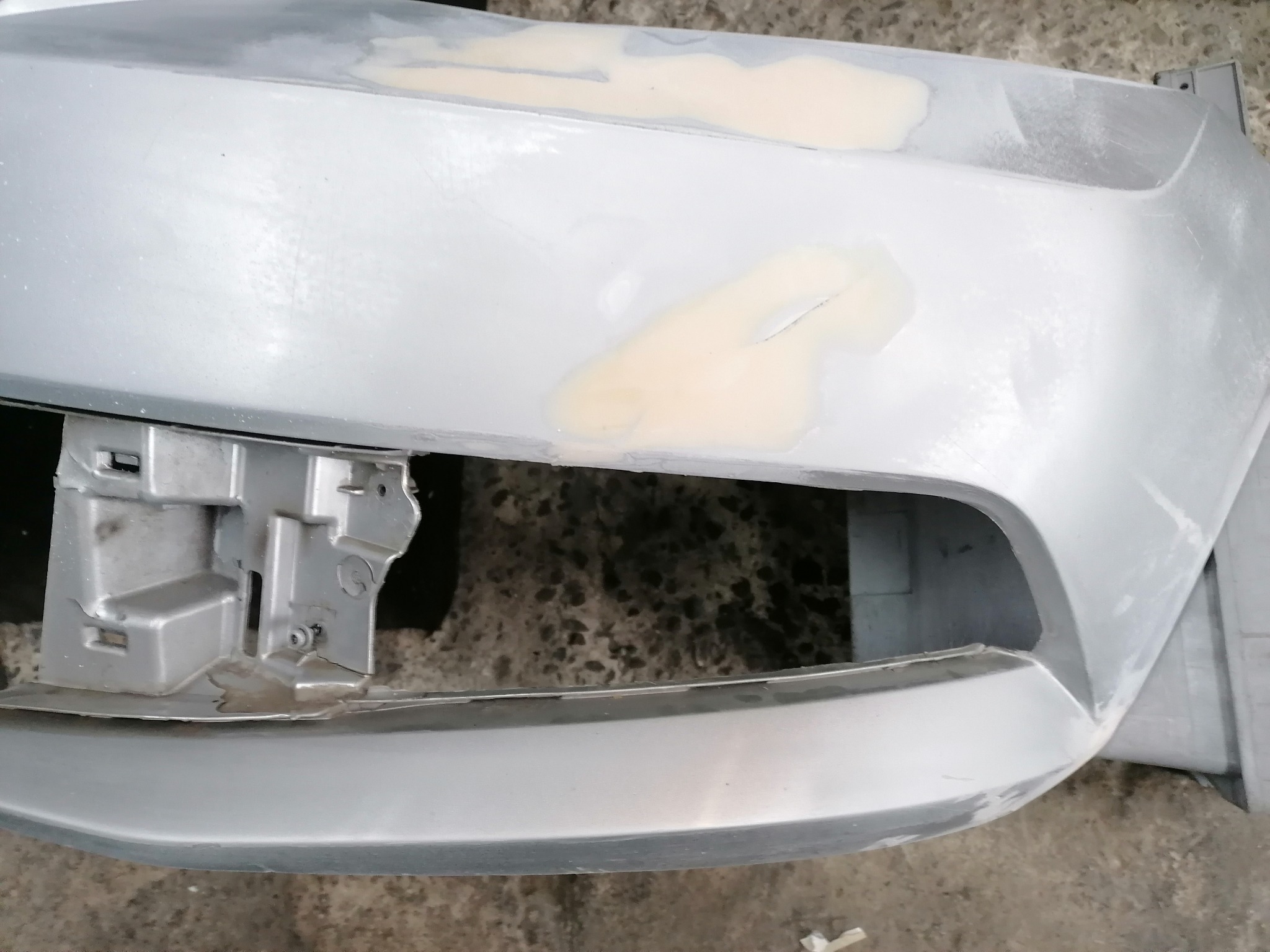 Szary zdemontowany zderzak samochodowy z widocznymi miejscami wypełnionymi szpachlą przed lakierowaniem, ukazujący proces naprawy karoserii.