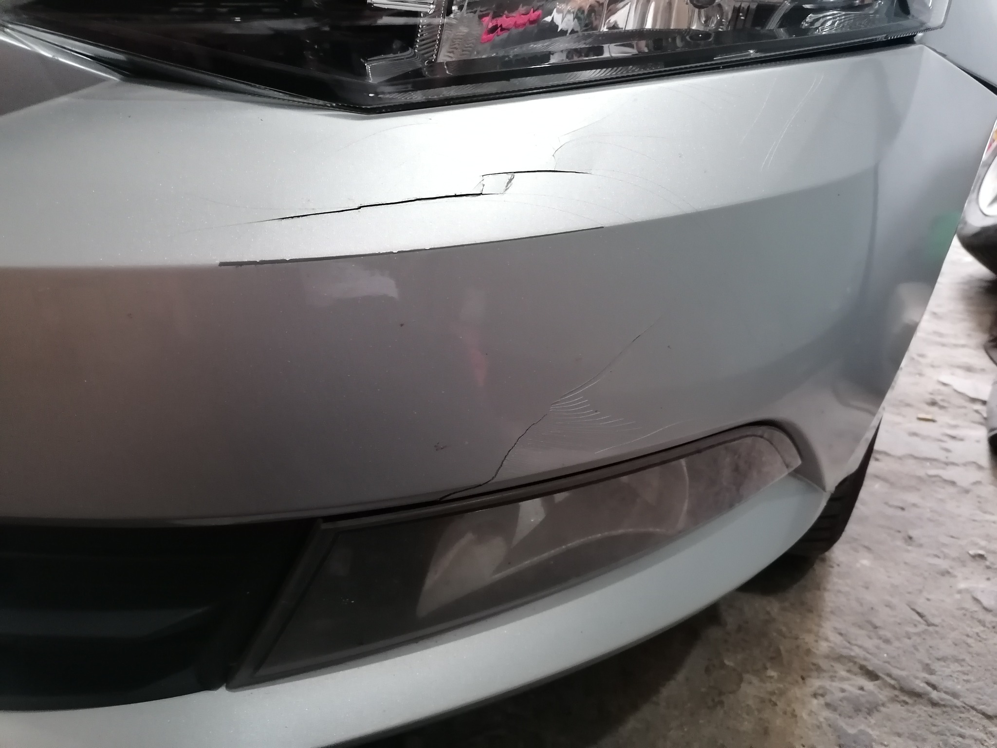 Uszkodzony, srebrny przedni zderzak samochodu z widocznymi pęknięciami i zarysowaniami lakieru, widok z bliska.