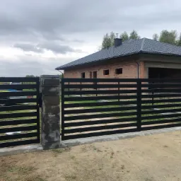 Nowoczesna, pozioma brama wjazdowa z czarnych, metalowych profili, osadzona na szarych, betonowych słupkach, na tle niedokończonego domu z cegły i pochmurnego nieba.