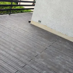 Szara deska kompozytowa na tarasie, widoczne łączenia desek, fragment ściany z gniazdami elektrycznymi i balustrada w tle.